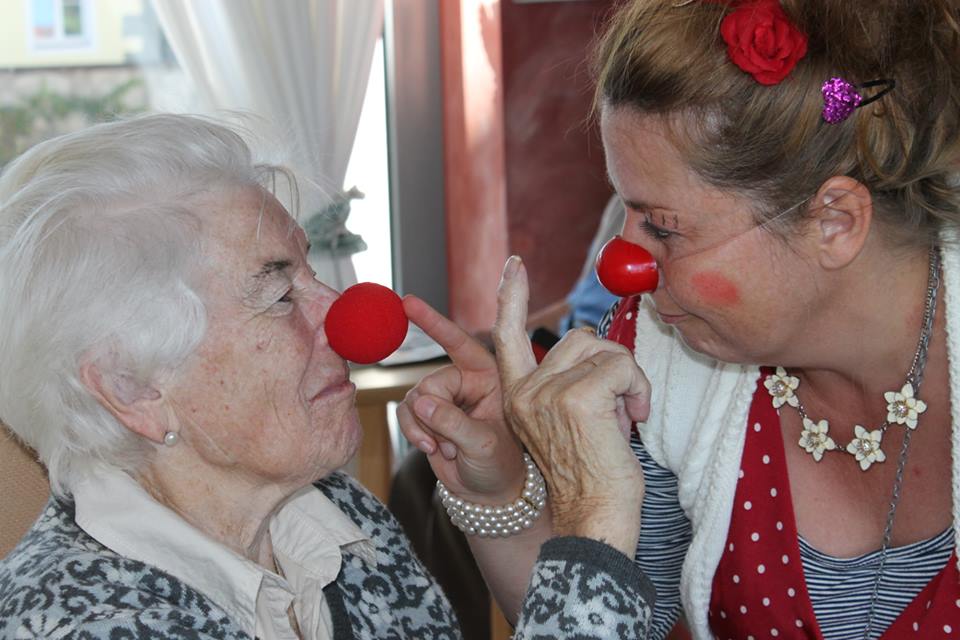Ältere Person und erwachsene Person mit roter Clownsnase sitzen sich nah gegenüber und berühren sich mit den Nasen, die erwachsene Person hebt einen Finger zwischen ihnen.