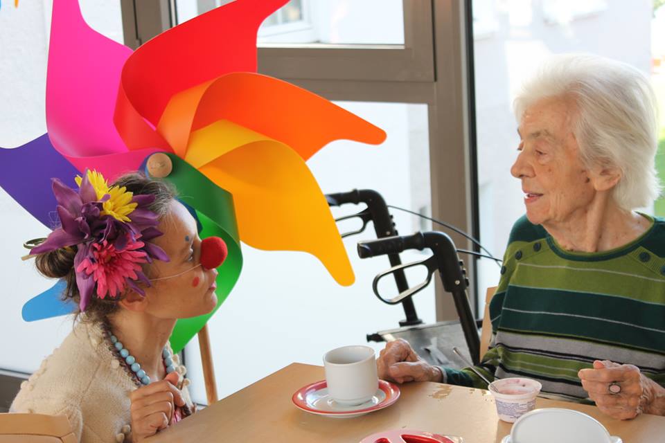 Erwachsene Person mit roter Clownsnase sitzt an einem Tisch gegenüber einer älteren Person, beide haben Tassen vor sich, im Hintergrund ein Rollator und ein buntes Windrad.