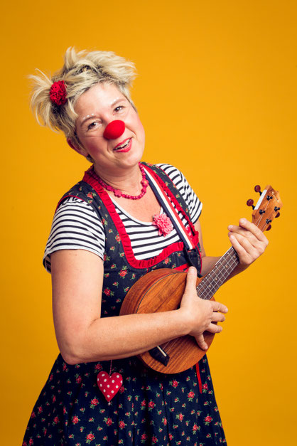 Erwachsene Person mit roter Clownsnase spielt eine Ukulele vor gelbem Hintergrund und blickt lächelnd in die Kamera.