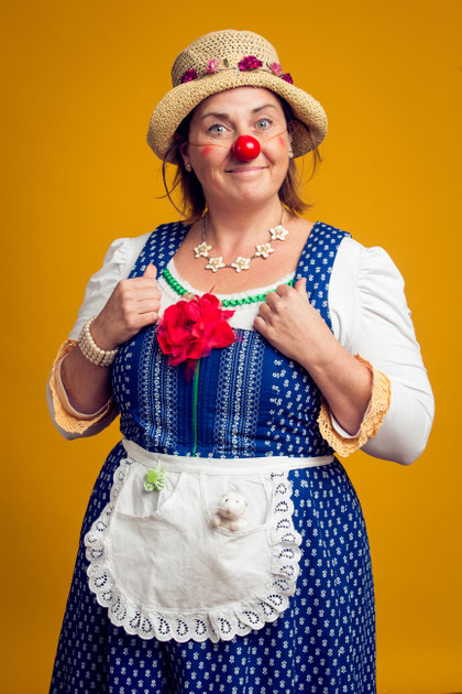 Erwachsene Person mit roter Clownsnase trägt ein blau-weißes Dirndl mit Schürze und Strohhut und steht vor gelbem Hintergrund.