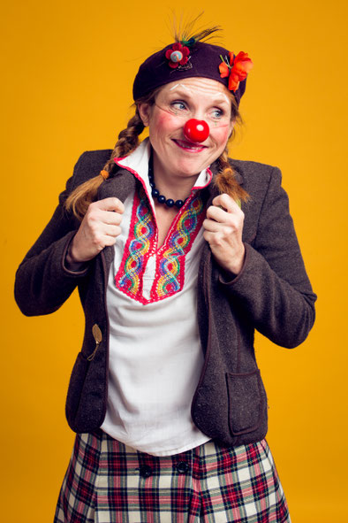 Erwachsene Person mit roter Clownsnase, geflochtenen Zöpfen und brauner Jacke steht vor gelbem Hintergrund und hält die Jackenkanten mit beiden Händen.