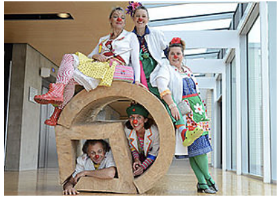 Fünf erwachsene Personen mit roten Clownsnasen posieren in einem hellen Innenraum um und auf einer großen, liegenden Skulptur