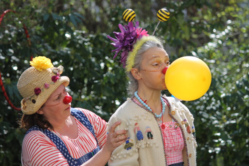 Zwei erwachsene Personen mit roten Clownsnasen stehen im Freien, eine bläst einen gelben Ballon auf, die andere berührt ihre Schulter.