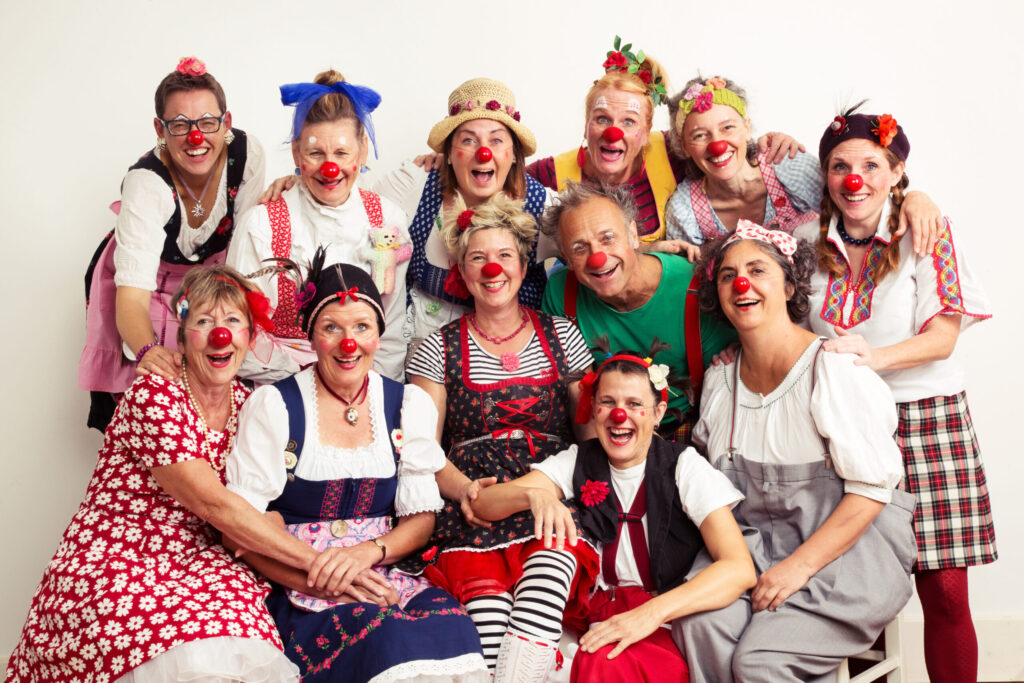 Gruppe von etwa 14 erwachsenen Personen mit roten Clownsnasen in unterschiedlichen Kostümen, dicht beieinander sitzend und stehend vor hellem Hintergrund.