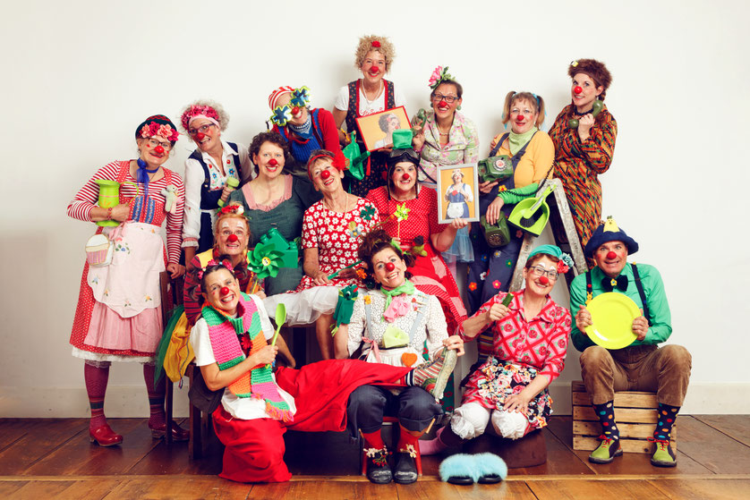 Gruppe von etwa 16 erwachsenen Personen mit roten Clownsnasen in bunten Kostümen, sitzend und stehend vor hellem Hintergrund, einige halten Requisiten wie Bilder, Küchenutensilien oder Instrumente.
