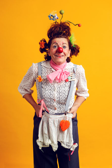 Erwachsene Person mit roter Clownsnase, hochgesteckten Haaren mit dekorativen Antennen, Bluse und Hosenträgern steht vor gelbem Hintergrund.