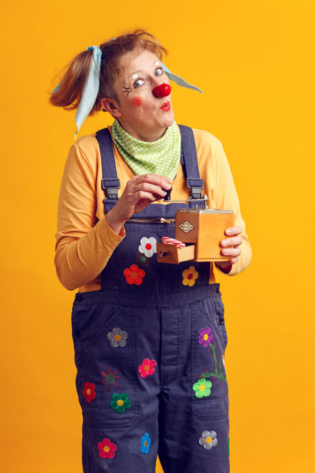 Erwachsene Person mit roter Clownsnase trägt eine Latzhose mit Blumenapplikationen und hält eine kleine Spieluhr in den Händen, vor gelbem Hintergrund.