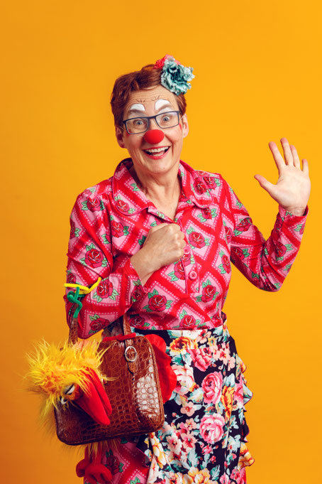 Erwachsene Person mit roter Clownsnase steht links vor gelbem Hintergrund und hält einen roten Koffer vor dem Körper.