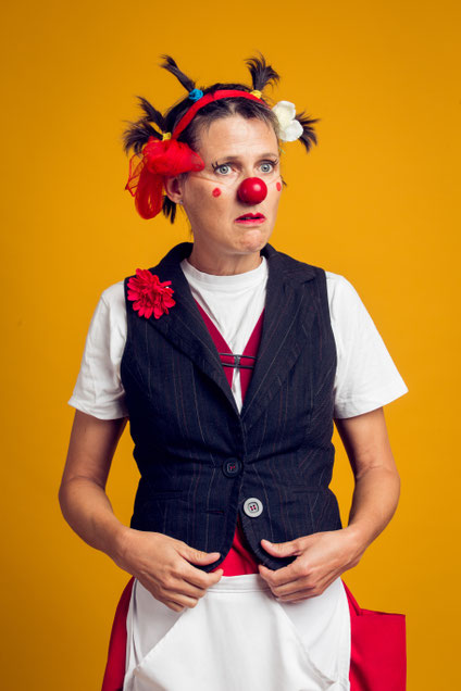 Erwachsene Person mit roter Clownsnase trägt ein weißes T-Shirt und eine schwarze Weste und steht vor gelbem Hintergrund.