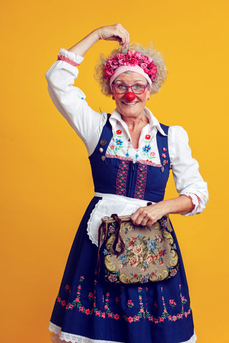 Erwachsene Person mit roter Clownsnase in traditionell wirkender Kleidung steht vor gelbem Hintergrund und hält eine gemusterte Tasche in der Hand.