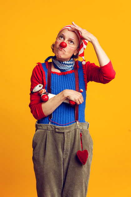 Erwachsene Person mit roter Clownsnase trägt ein rotes Oberteil, blaue Latzhose und eine gestreifte Mütze und steht vor gelbem Hintergrund.