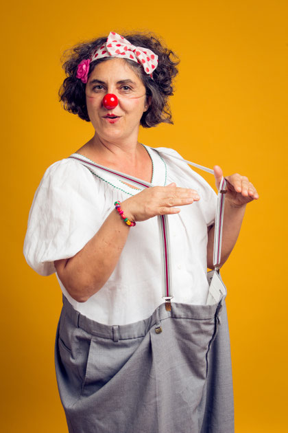 Erwachsene Person mit roter Clownsnase, weißer Bluse und Hosenträgern steht vor gelbem Hintergrund und hält die Hände waagrecht vor dem Körper.