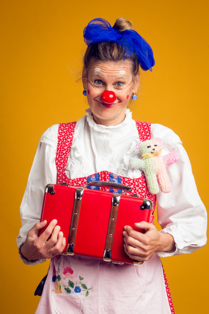 Erwachsene Person mit roter Clownsnase hält einen roten Koffer vor der Brust, trägt ein buntes Kostüm und steht vor gelbem Hintergrund.
