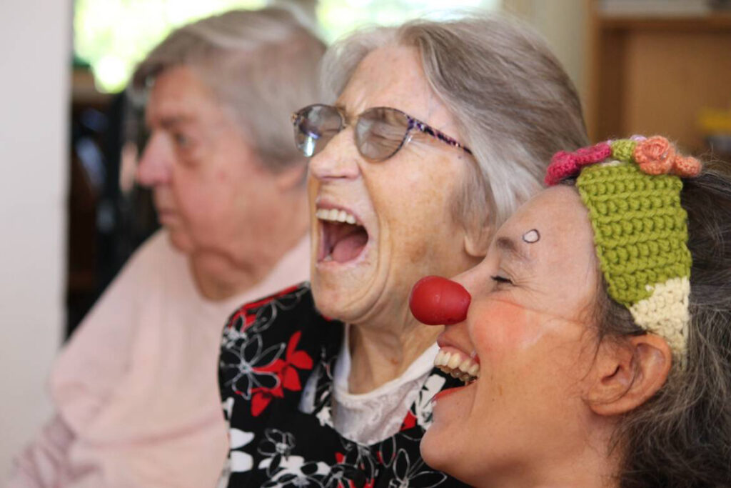 Zwei ältere erwachsene Personen sitzen nebeneinander in einem Innenraum und lachen, eine davon trägt eine rote Clownsnase und eine gehäkelte Kopfbedeckung.